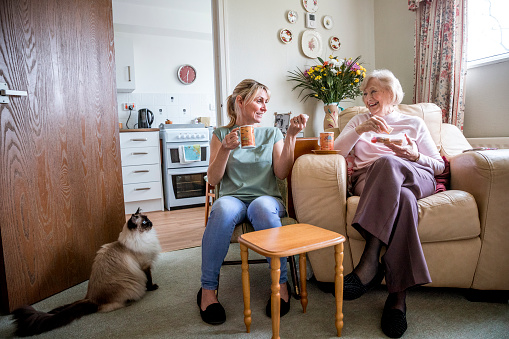 Senior woman sitting in her armchair listening to a younger woman talking. Both look happy as they enjoy a drink and snack together