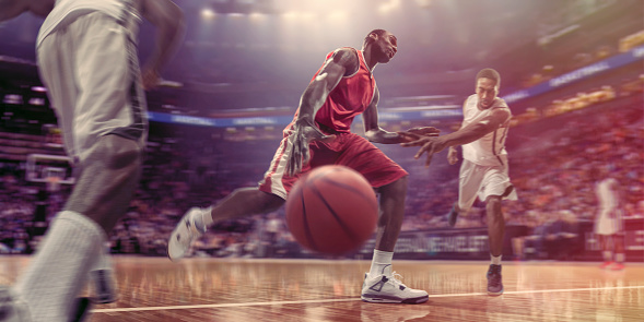 A close up, wide angle, low viewpoint image of a professional male basketball player, dressed in red basketball uniform running and dribbling a basketball. The basketball player is moving into a challenge by an opposition player who is reaching for the ball. The action occurs in an generic indoor arena.