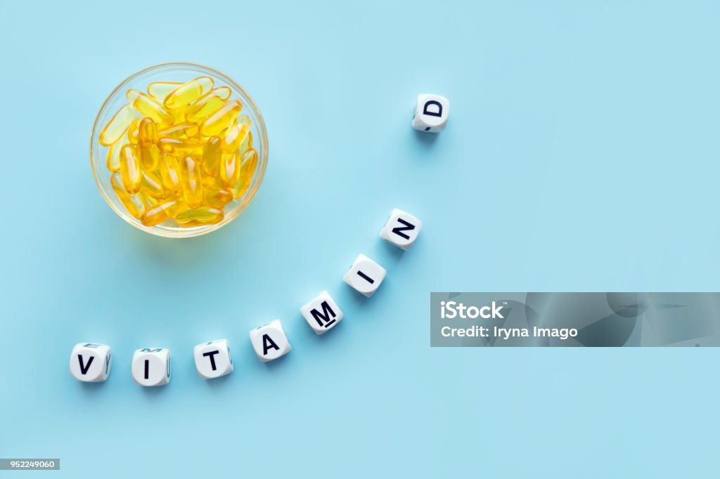 Yellow capsules in the round glass bowl and the word vitamin D from white cubes with letters on a blue background Yellow capsules in the round glass bowl and the word vitamin D from white cubes with letters on a blue background. VITAMIN D word for healthy and medical concept. Sunshine vitamin health benefits Vitamin D Stock Photo