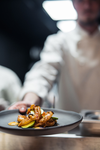 Restaurant Chef cook holding dish with shrimps and avocado. The plate with meal is on foreground and in focus. Cook is on the background and blurred.