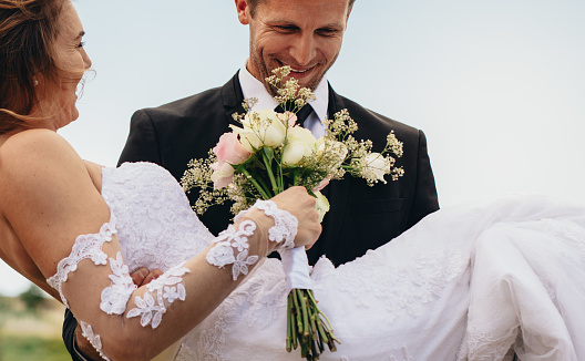 Happy groom carrying beautiful bride with flowers in hand. Newlywed couple with bouquet outdoors on their wedding day.