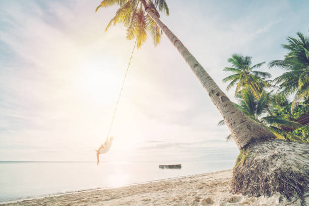 istock Young woman playing on beach, swing rope on palm tree 952366292