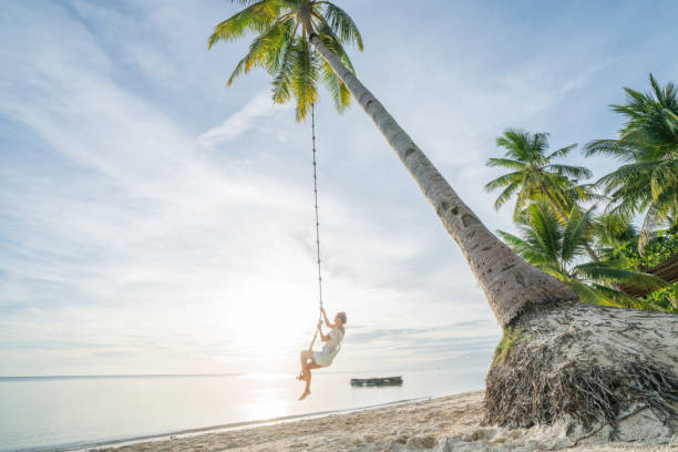 istock Young woman playing on beach, swing rope on palm tree 952366676