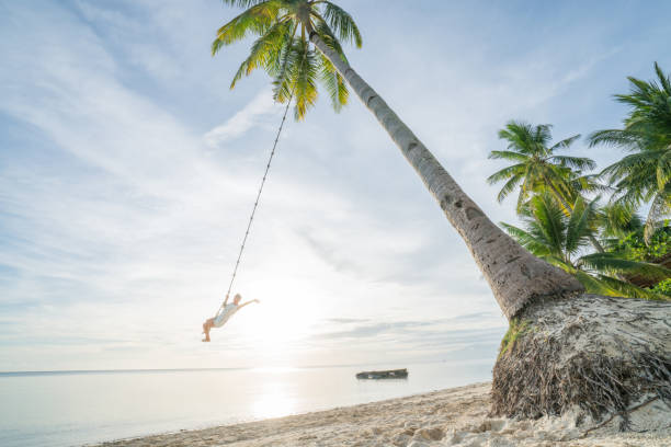 istock Young woman playing on beach, swing rope on palm tree 952366902
