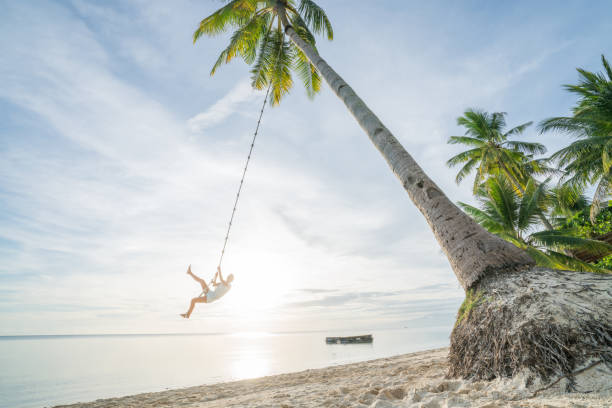 istock Young woman playing on beach, swing rope on palm tree 952366906
