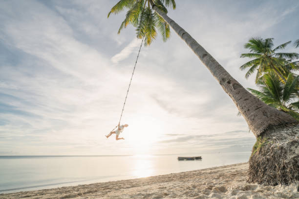 istock Young woman playing on beach, swing rope on palm tree 952367084