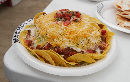 Layers of nachos on a Styrofoam plate piled with hotdogs, salsa, jalapeno and topped with shredded cheese and sliced tomatoes.