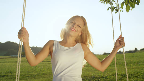 istock LENS FLARE PORTRAIT: Smiling sunlit woman in white sundress swinging under tree. 952437240