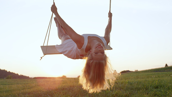 LENS FLARE CLOSE UP: Illuminated in bright sunshine, young woman sways in nature and leans back on rope swing. Summer evening sunrays shining down on girl having fun on lonely swing in rural setting.