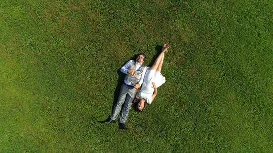 AERIAL TOP DOWN: Happy young couple laying in grass on summer day. Man and wife newlyweds relaxing in sunny park after wedding. Young man and pregnant woman lying and holding hands, expecting a baby