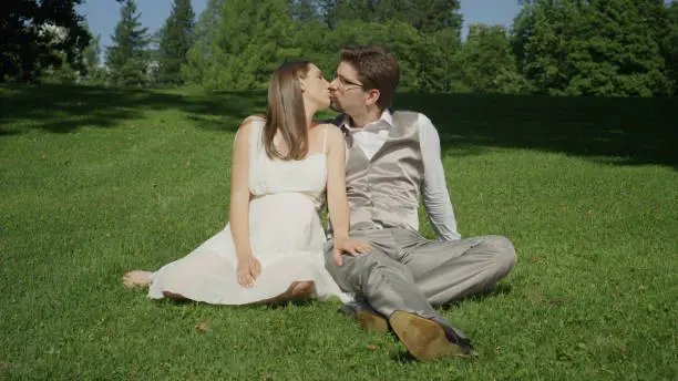 Beautiful young couple sit on grass in a beautiful sunlit park playfully kissing. Expecting parents share some tenderness on a warm sunny day. Newlyweds having a wonderful time outdoors in summer.