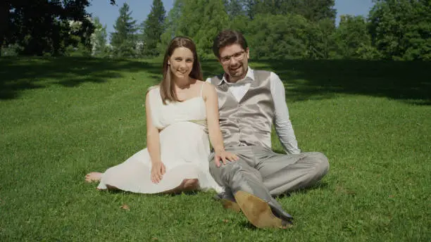 Young couple sitting on grass in a beautiful sunlit park posing for the camera. Expecting parents share some tenderness on a warm sunny day. Newlyweds having a wonderful time outdoors in summer.