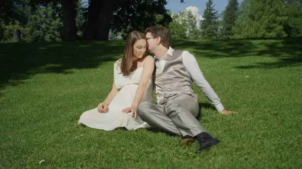 Loving couple sitting in a green meadow on beautiful sunny day being romantic. Parents to be enjoying day out in the sun. Affectionate husband giving a kiss on the head to his beautiful pregnant wife.