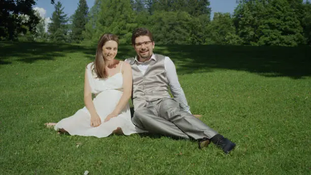 PORTRAIT Excited pregnant couple sitting on grass in beautiful green meadow smiling at camera. Young parents to be relaxing on a sunny summer day. Newlyweds enjoying peace and quiet after the wedding.