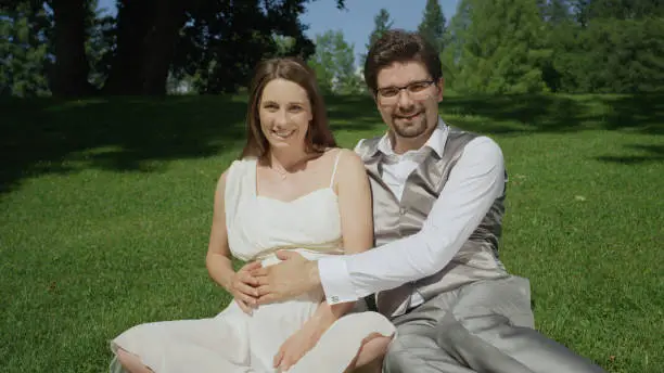 PORTRAIT: Smiling couple sitting on grassy floor in a beautiful garden on a sunny day gently caressing her pregnant belly. Newlyweds sharing an intimate moment. Expecting parents enjoying pregnancy.