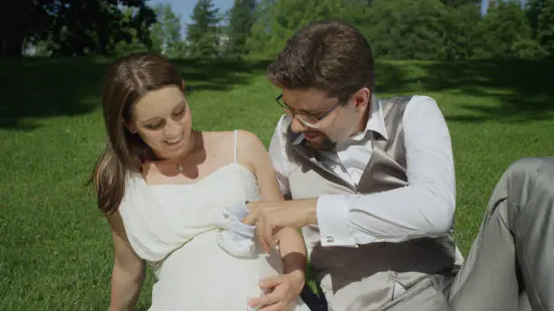 CLOSE UP: Excited father to be playfully touching wife's pregnant belly with tiny blue baby shoes. Expecting parents relaxing in a beautiful green garden on a sunny day. Newlyweds unwinding in park.