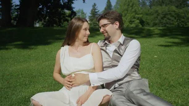 CLOSE UP: Beautiful young couple in love caressing her baby bump waiting for baby to kick. Husband and wife relaxing in a tranquil green park on a sunny summer day. Parents to be enjoying pregnancy.