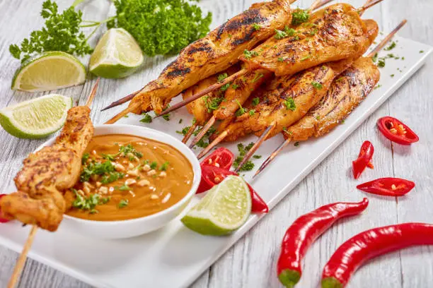 grilled spicy Chicken satay on skewers on a white rectangular plate with peanut sauce. red chili peppers, lime slices and parsley on table, view from above, close up