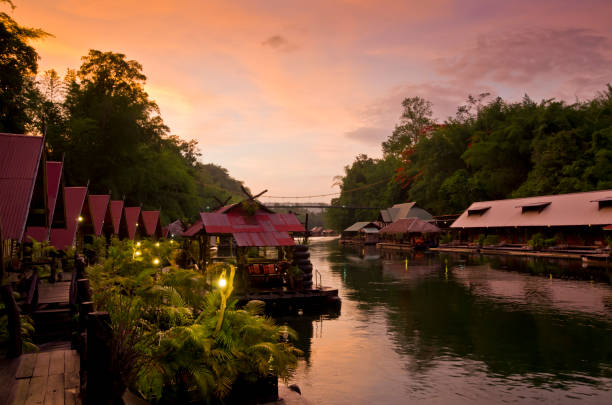 The river Kwai or Kwai, also known as the river Kwai Noi at sunset with floating rafts either side of the river. In the middle distance is a footbridge
