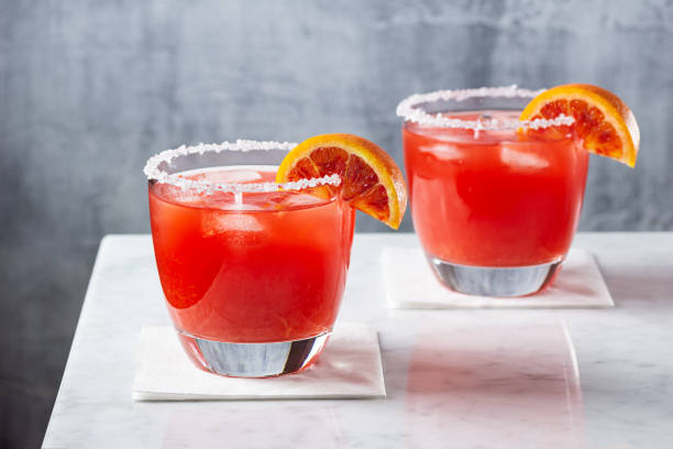 Blood Orange Margaritas on the Rocks with Salted Rims on Marble Bar Two blood orange margaritas served on the rocks in glasses with salted rims. Blood orange wedges garnish the cocktails sitting on napkins on a white marble bar. The minimalist gray background has space for copy. tequila drink stock pictures, royalty-free photos & images