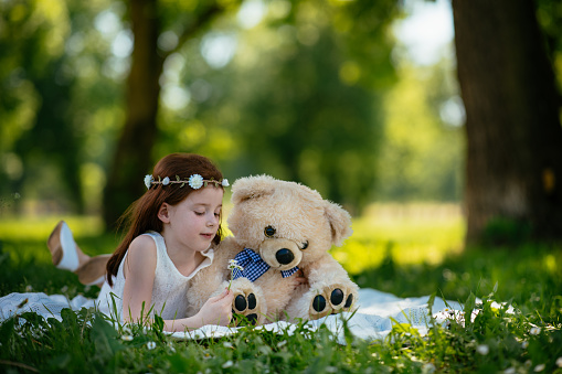 a little girl with curly blond hair sits on a stump in the courtyard of the house and tightly hugs her favorite soft toy teddy bear