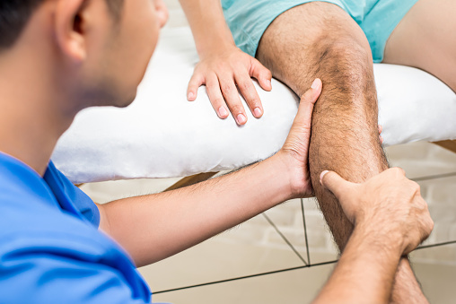 Doctor (therapist) treating male patient leg while sitting on the bed in clinic - physical therapy concept