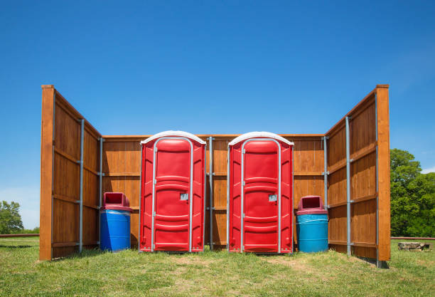 Two red portable restrooms with a wood fence around them in a park Two red portable restrooms and trash cans with a wood fence around them in a park. Trees and blue sky background. portable toilet stock pictures, royalty-free photos & images