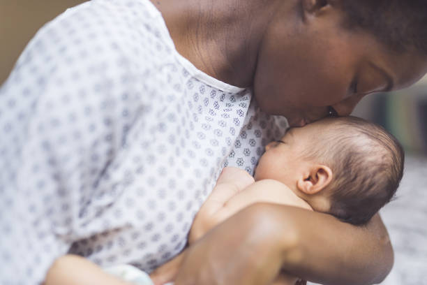 New Mom Holds Her Infant to Her Chest A beautiful young African American mother in a hospital gown gently holds her infant in her arms and smiles down at her. The baby's eyes are closed. newborn stock pictures, royalty-free photos & images