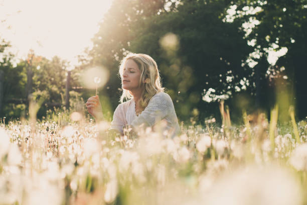 red-haired teenage girl, in swimsuit and shorts, sits on grass, blows on dandelion, winces from an allergy to fluff, on sunny summer day