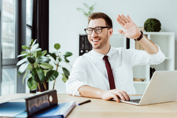 istock cheerful businessman waving and using laptop at workplace 952747588