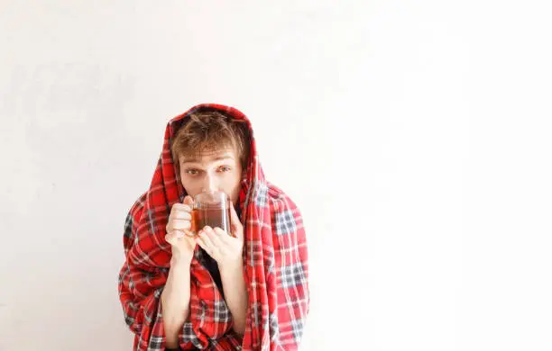 Man cold, has got sick. It was wrapped up in a red plaid and has drinking tea, against background of a white wall. Copy space