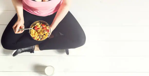 Sports girl healthy breakfast. Girl sits on a wooden floor in leggings, a pink undershirt and sneakers. Panorama. Copy space