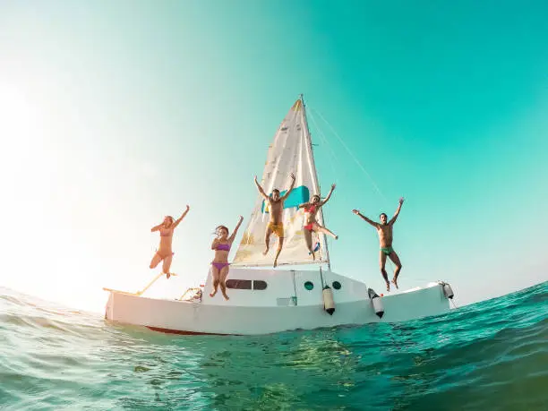 Photo of Happy crazy friends diving from sailing boat into the sea - Young people jumping inside ocean in summer vacation - Main focus on center guys - Travel and fun concept - Fisheye lens distortion