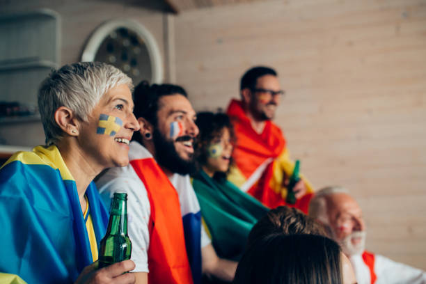 зрелая женщина, поддерживающая шведскую футбольную команду, смотрит игру с друзьями, поддерживающими другие сборные - german culture people women germany стоковые фото и изображения