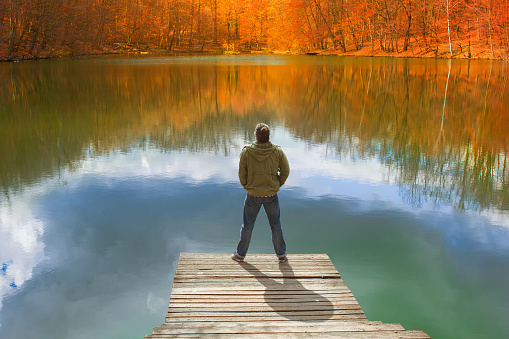 Lonely man standing on the edge of the small wooden pier looking at the lake and colorful forest on the other side