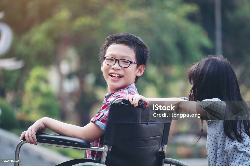 Ragazzo felice in sedia a rotelle con ragazza prova a guidare una sedia a rotelle del sorriso del fratello, tempo di famiglia - Foto stock royalty-free di Bambino