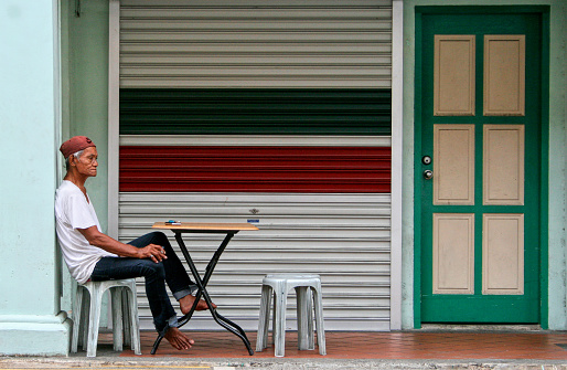 Little India, Singapore - June 7, 2008: An old man rests at an outdoor table in front of a building with closed shutter and door while he smokes a cigarette