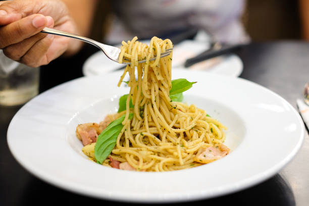 istock Woman's hand holding fork for eating spaghetti spicy bacon & ham with basil, tomato, black pepper, lettuce and paprika on white dish set on table for food background or texture. 953022142