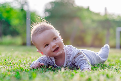 Cute funny laughing Caucasian baby boy learning to crawl, having fun playing on the lawn watching summer flowers in the garden during bright sunny day. Photo of smiling small 6 months old boy in green grass - he smiles and has fun while looking up. Happy young baby lying on tummy in nature.
