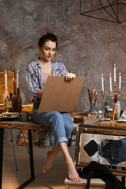 istock Attractive woman painter sitting on the table, drawing and smiling. Creative concept. Drawing supplies, artist brushes, canvas, candle, oil lamp 953039748