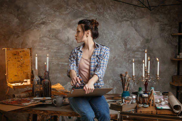 istock Woman painter sitting on the table, drawing and looking left. Creative concept. Drawing supplies, oil paints, artist brushes, canvas, candle 953039956