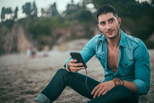 Young handsome man sitting at the beach and using phone