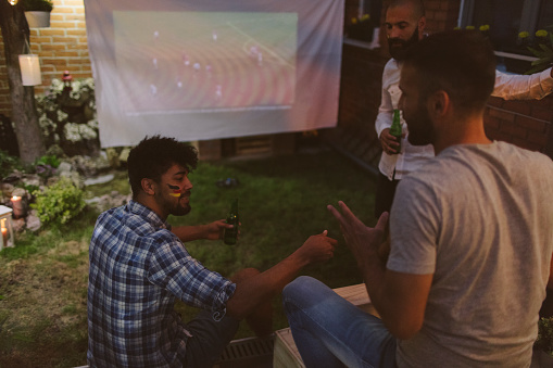 Friends watching soccer game in backyard on big screen and drinking beer. German national flag on face.
