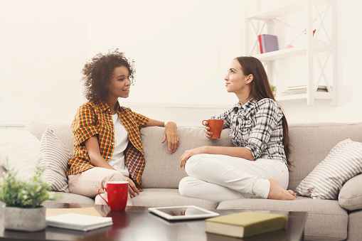 Two happy young female friends with coffee cups conversing in living room at home, chatting about their life and relations, gossip and slumber party concept, copy space