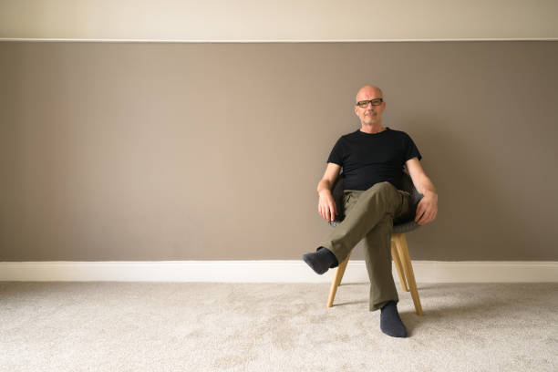 Moving in to new home DIY Mature man moving in to a new home sits on a chair in an empty room; walls painted, new carpet, ready for the furniture. legs crossed at knee stock pictures, royalty-free photos & images