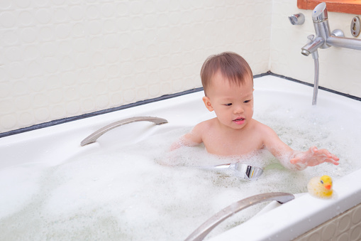 Cute little Asian 18 months / 1 year old toddler baby boy child having fun sitting in bathtub playing rubber duck toy and take a shower by himself in the bathroom indoor at home, baby care concept