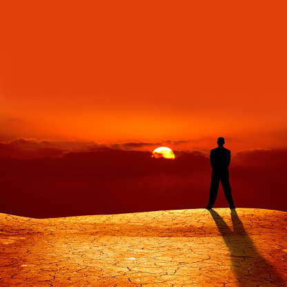 conceptual image of silhouetted businessman on cracked dry landscape watching sunset