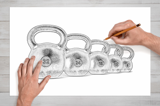 Male hands drawing several kettlebells of different weight with a pencil on white paper. Sports and motivation. Creating strong body. Art and strength.