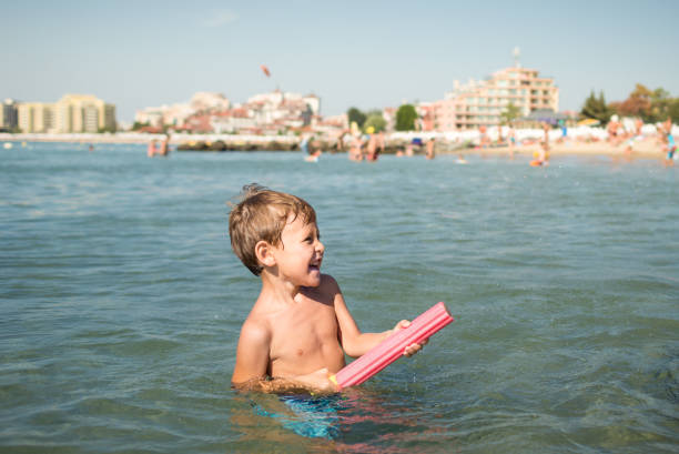 Joyful little boy shoots from a water-pistol at someone in the sea. Funny child plays in the water