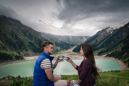 Young couple looks at each other and holds in hands traditional cups. Handsome man drinks tea with girlfriend. Majestic mountains with snowy tops, lake and dramatic cloudy sky.